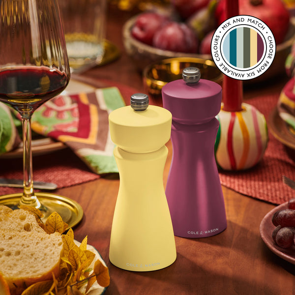 Two stylish Cole & Mason Kenton Salt & Pepper Mills—one in Turmeric yellow, one purple—stand on a festive table with wine, bread, and autumn decor. A circular label highlights six color options and their durable ceramic mechanism.