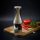 The Cole & Mason Kew Electronic 2-in-1 Duo Mill sits on a wooden cutting board, surrounded by a tomato, two red chili peppers, herbs, and garlic cloves. The grinder features adjustable settings for salt and pepper.