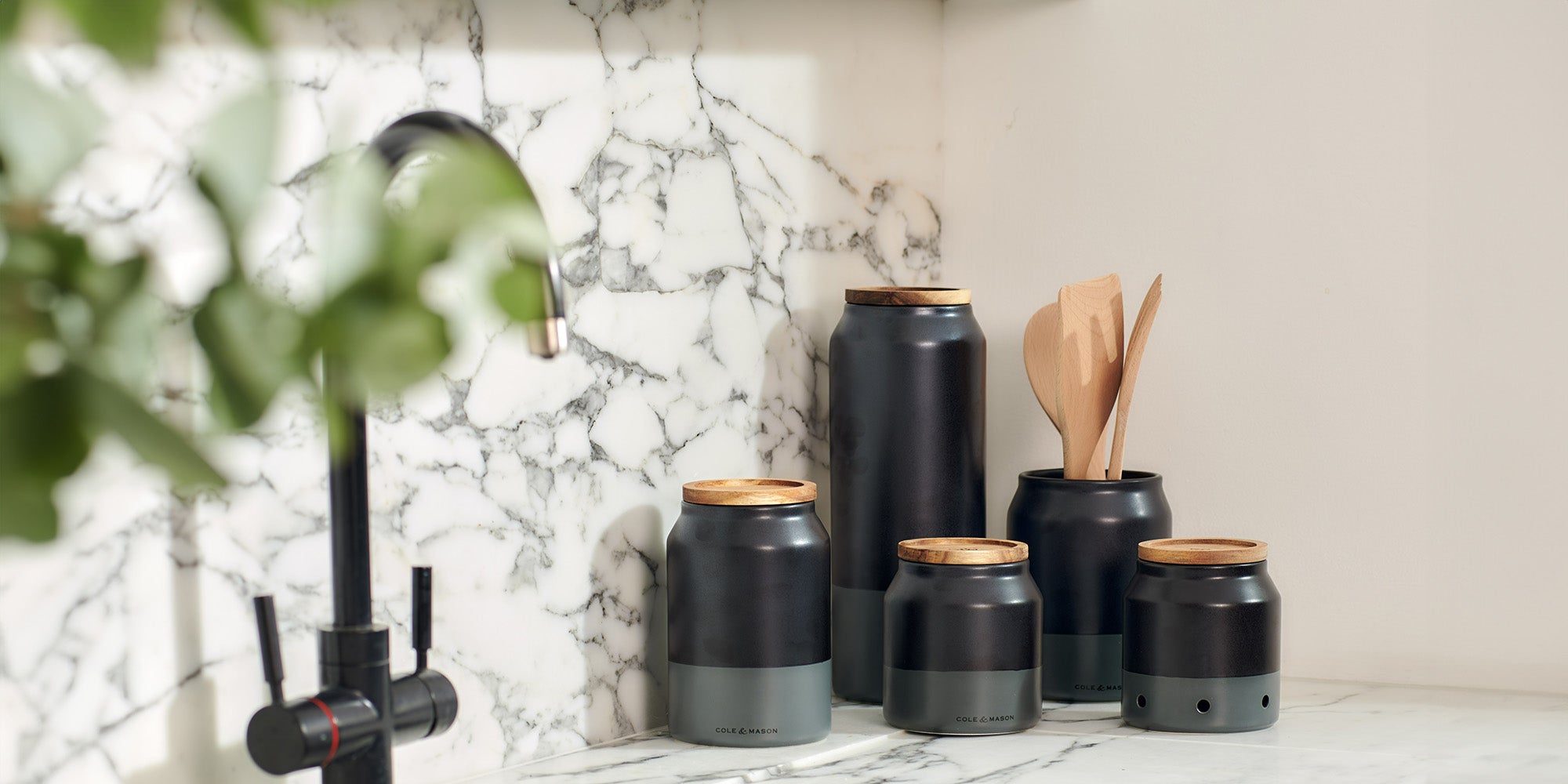 A set of black and gray kitchen canisters with wooden lids sits on a marble countertop near a sink; one jar holds wooden utensils and sunlight shines into the scene.