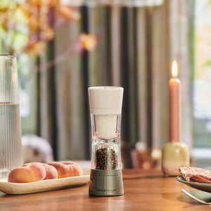 The Cole & Mason Lincoln 2-in-1 Duo Mill with adjustable grinding sits on a wooden table beside peaches, a tall glass of water, and a lit candle, while soft natural light streams through the curtains behind.