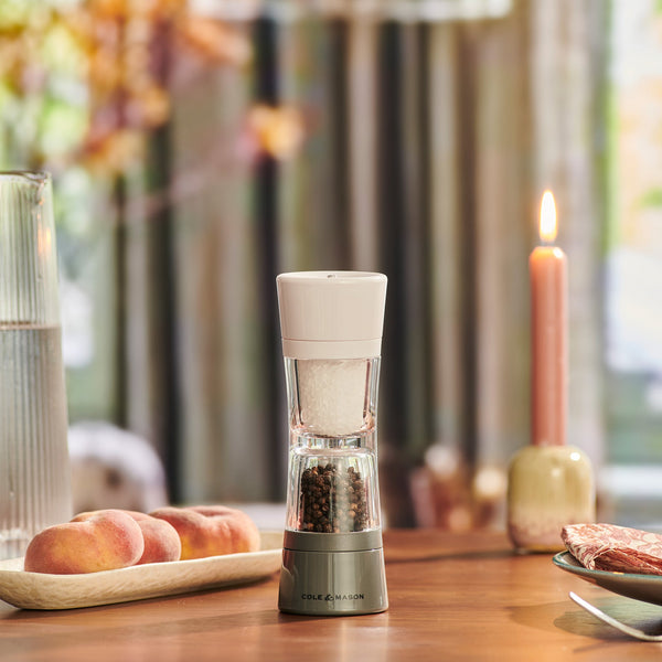 The Cole & Mason Lincoln 2-in-1 Duo Mill with adjustable grinding sits on a wooden table beside peaches, a tall glass of water, and a lit candle, while soft natural light streams through the curtains behind.