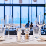 #130mm Two clear Cole & Mason London Acrylic Salt & Pepper Shakers are on a set restaurant table with wine and water glasses and plates, positioned by large windows overlooking a blurred cityscape.