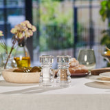 #130mm Two clear Cole & Mason London Acrylic Salt & Pepper Shakers rest on a white tablecloth among plates, glasses, bread, and flowers, with a softly blurred outdoor background.