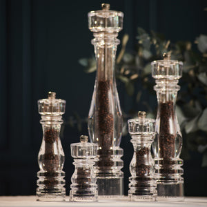 The Cole & Mason London Acrylic Salt & Pepper Mills, filled with dark peppercorns and available in various heights, are displayed on a light surface with a blurred green background—ideal for any modern kitchen.