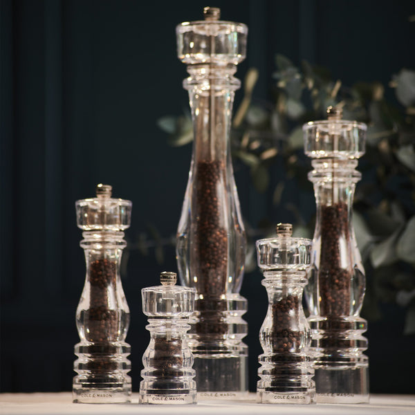 The Cole & Mason London Acrylic Salt & Pepper Mills, filled with dark peppercorns and available in various heights, are displayed on a light surface with a blurred green background—ideal for any modern kitchen.