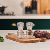 #130mm #shakerset Two transparent Cole & Mason London Acrylic Salt & Pepper Shakers sit on a wooden cutting board beside fresh artichokes on a kitchen counter, with a stove and utensils softly blurred in the background.