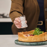 #86mm 
A person in a brown jacket seasons a savory pie topped with fresh greens, using Cole & Mason London Acrylic Salt & Pepper Shakers over the dish on a marble surface.