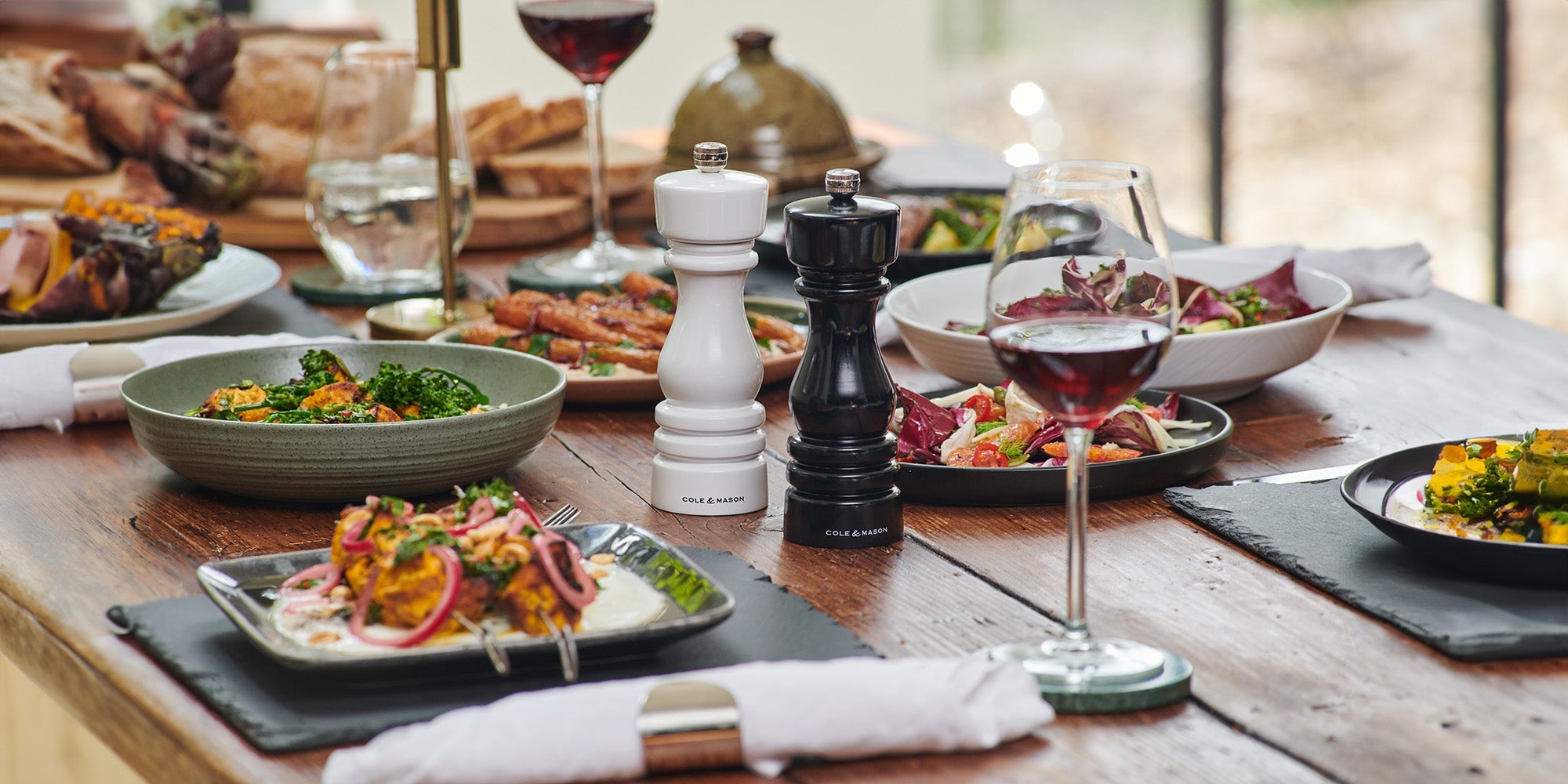 A wooden dining table set with plates of colorful salads, roasted vegetables, cutlery, wine glasses filled with red wine, white and black pepper mills, and napkins, creating an elegant meal setting.