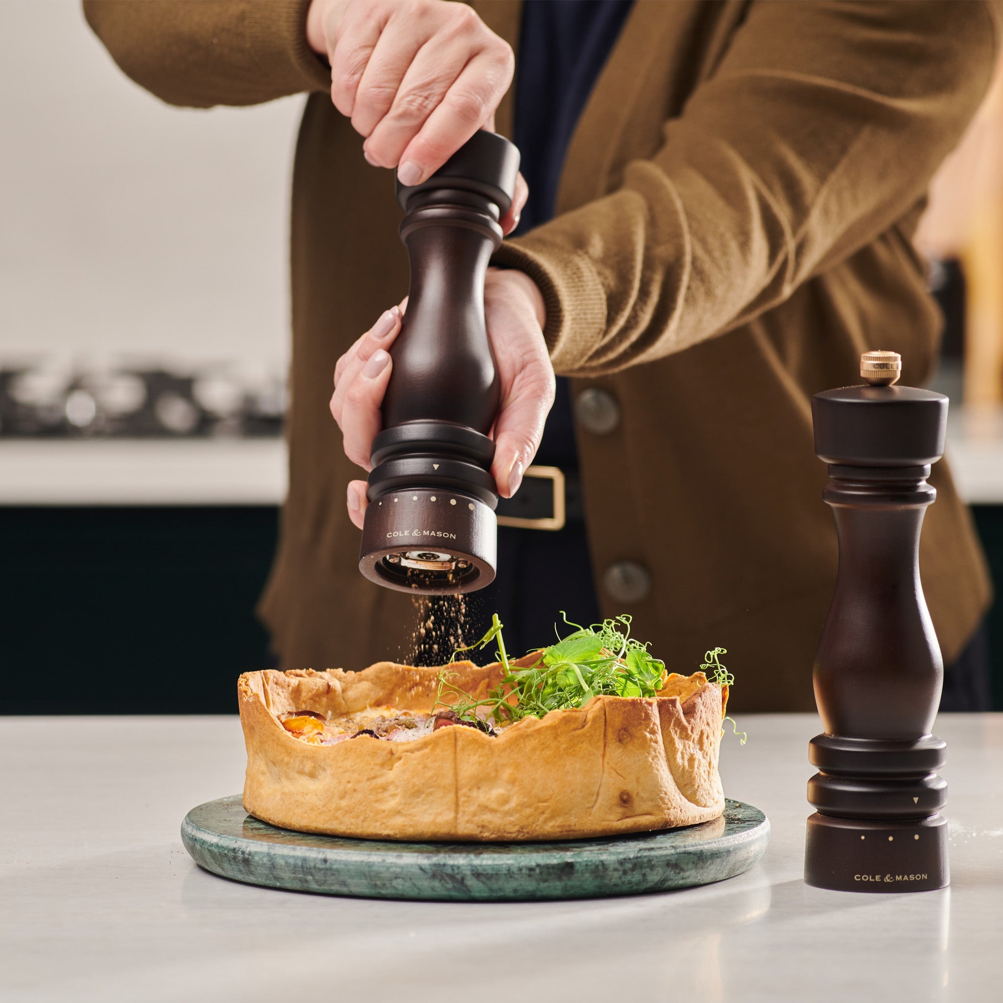 A person grinds pepper onto a savory pie topped with greens, using a wooden pepper mill. Another pepper mill stands nearby on the white countertop. The scene is set in a modern kitchen.
