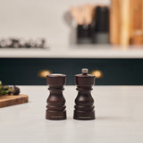 The Cole & Mason London Chocolate Wood Salt Shaker & Pepper Mill Set sits side by side on a white kitchen countertop, crafted from natural beech wood with a rich dark finish. Blurred utensils and kitchen items are visible in the background.