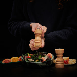 A person in a dark sweater seasons salad with the Cole & Mason London Natural Beech Salt Shaker & Pepper Mill Set, next to sliced citrus, bread, a cherry tomato, and another matching pepper grinder on the table.