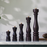 Five Cole & Mason London Chocolate Wood Salt & Pepper Mills of varying heights stand in ascending order on a marble countertop, backed by a white wall with soft natural light and plant shadows. Premium grinding technology ensures effortless use.