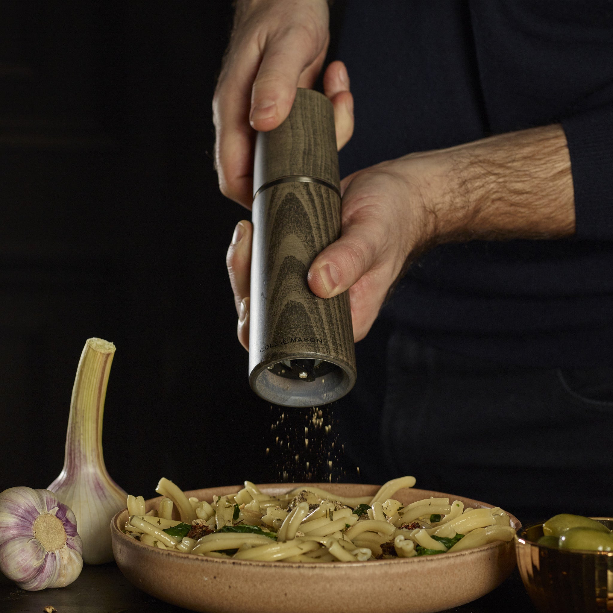 A person grinds black pepper onto a plate of pasta, garnished with herbs and nuts. A bulb of garlic and a bowl of olives are visible nearby on the table.