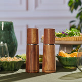 Two Marlow Natural Acacia Salt & Pepper Mills by Cole & Mason UK, featuring adjustable grinding mechanisms, sit on a green table with bowls of food and glasses, delivering precise seasoning in a stylish dining setting.