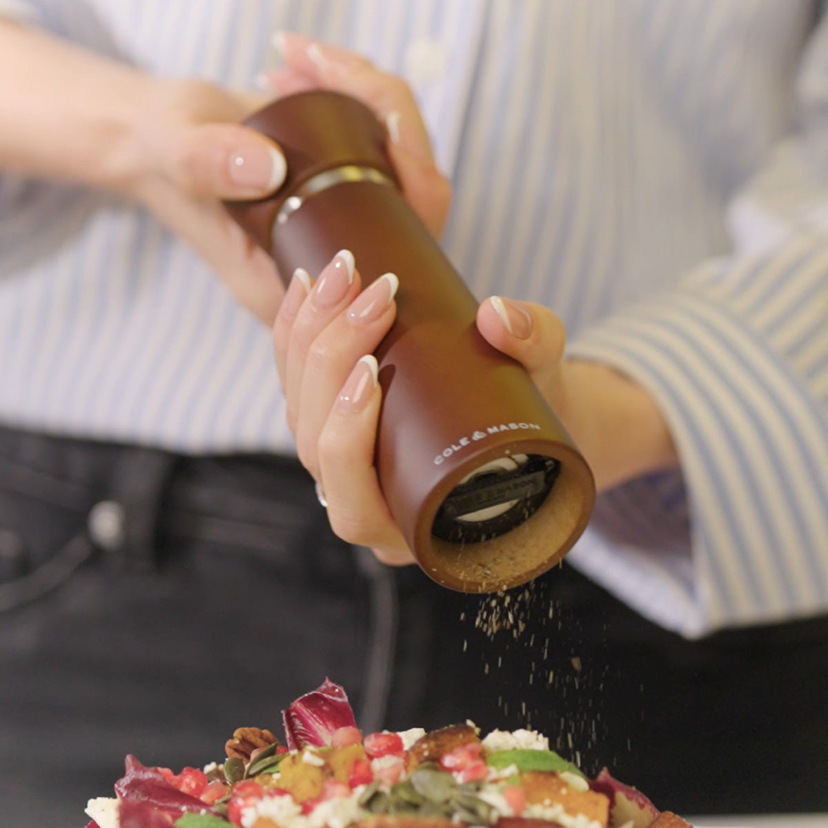 A person in a striped shirt uses a brown Cole & Mason grinder to sprinkle spices over a colorful salad with greens, seeds, and pomegranate seeds.