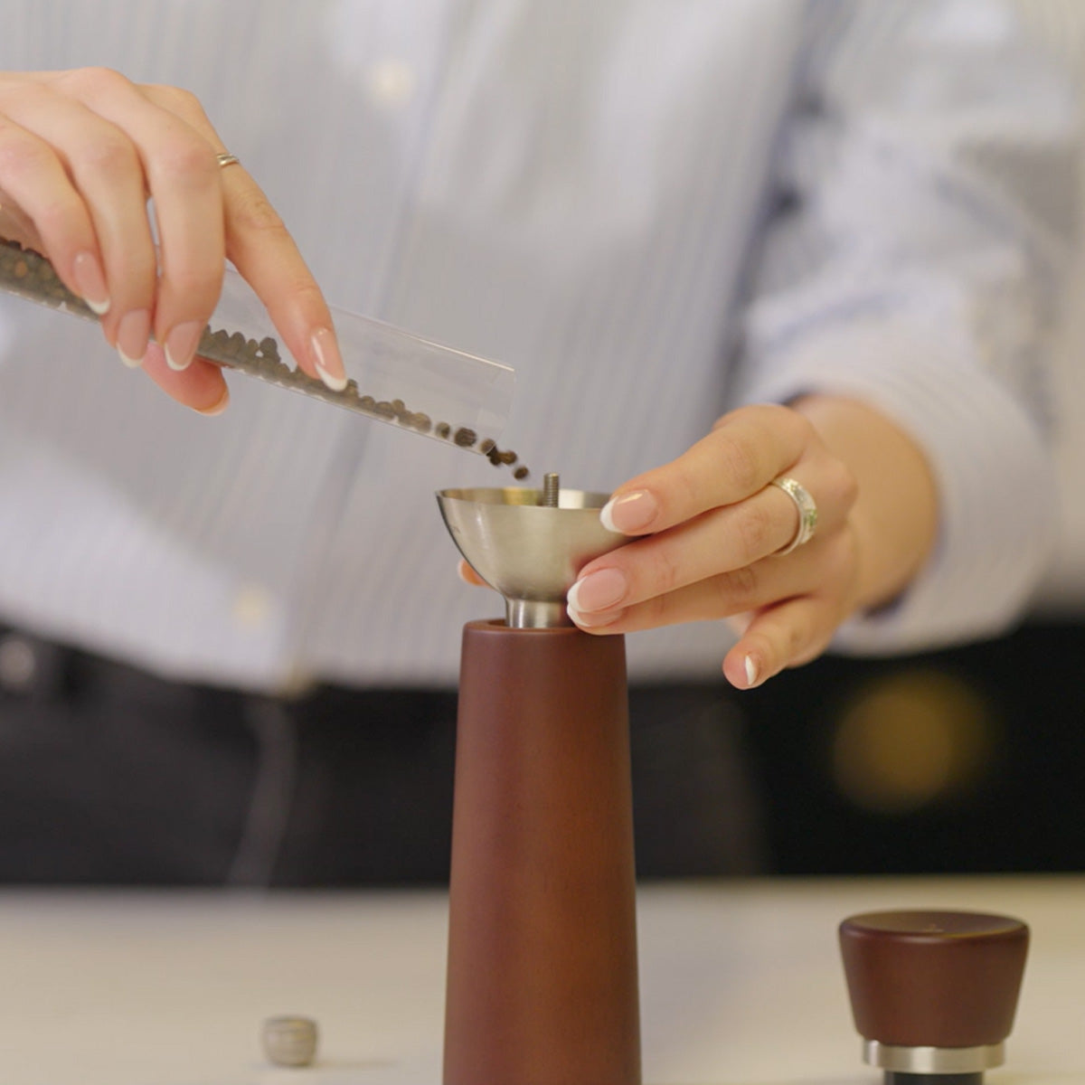 A person pours whole black peppercorns from a small plastic container into the top of a brown pepper grinder, preparing to refill it. The person is wearing a light blue striped shirt.