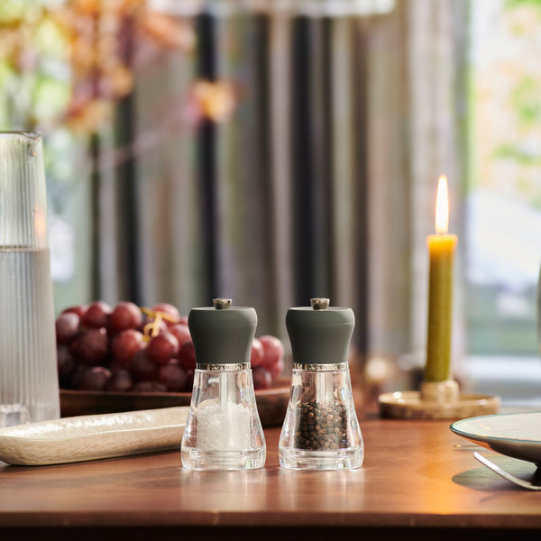 The Cole & Mason Napoli Salt & Pepper Mill Set, with clear bodies and grey tops, sits on a wooden table ready for a meal, surrounded by grapes, a glass of water, a lit candle, and bathed in soft natural light.
