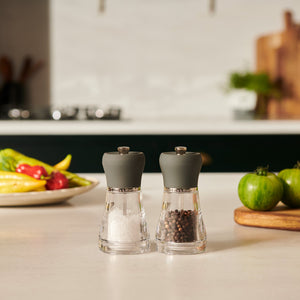 The Cole & Mason Napoli Salt & Pepper Mill Set, featuring glass bodies and gray tops, sits elegantly on a kitchen counter. With high-quality grinding mechanisms, these mills ensure perfect seasoning in any modern kitchen setting.