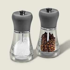The Cole & Mason Napoli Salt & Pepper Mill Set features clear glass bodies with gray tops, elegant design, and premium grinders. Filled with white salt crystals and whole brown peppercorns, they stand side by side on a light background.