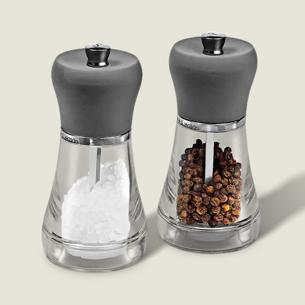The Cole & Mason Napoli Salt & Pepper Mill Set features clear glass bodies with gray tops, elegant design, and premium grinders. Filled with white salt crystals and whole brown peppercorns, they stand side by side on a light background.