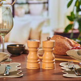 The Cole & Mason London Natural Beech Salt Shaker & Pepper Mill Set is displayed on a dining table set with glassware, utensils, napkins, bread, and greenery in the background.