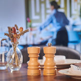 A Cole & Mason London Natural Beech Salt Shaker & Pepper Mill Set rests on a restaurant table with plates, napkins, and dried flowers in a glass vase, while a blurred figure works in the background.