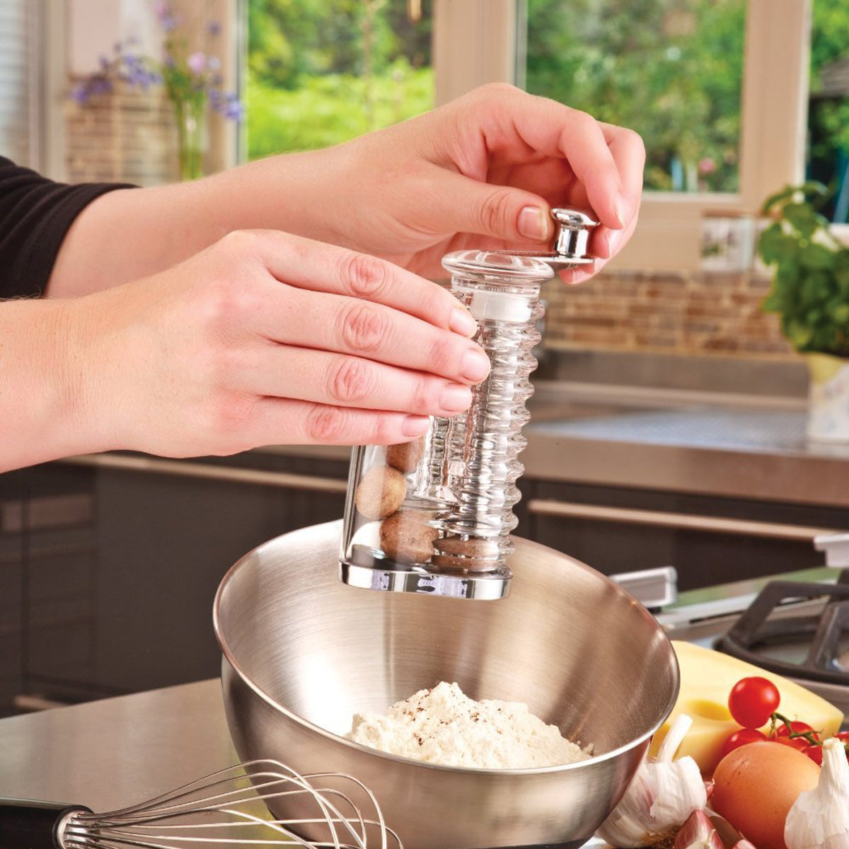A person grinds whole nutmeg into a stainless steel mixing bowl filled with flour in a bright kitchen, with a whisk and various fresh ingredients nearby.