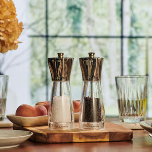 The Cole & Mason UK Rye Salt & Pepper Mill Set 160mm sits on a wooden board with glasses, plates, and peaches nearby, while a blurred window provides the background.