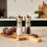 The Cole & Mason UK Rye Salt & Pepper Mill Set 160mm features two clear grinders with silver tops and durable ceramic mechanisms, shown on a wooden cutting board with figs on a light kitchen countertop.