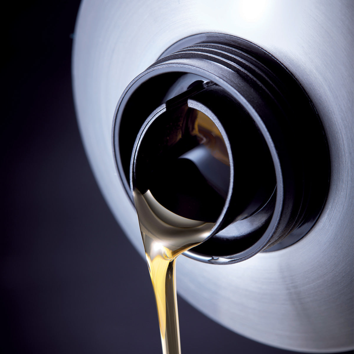 A close-up of motor oil being poured from a silver container, with the golden liquid flowing smoothly from the bottles black spout against a dark background.
