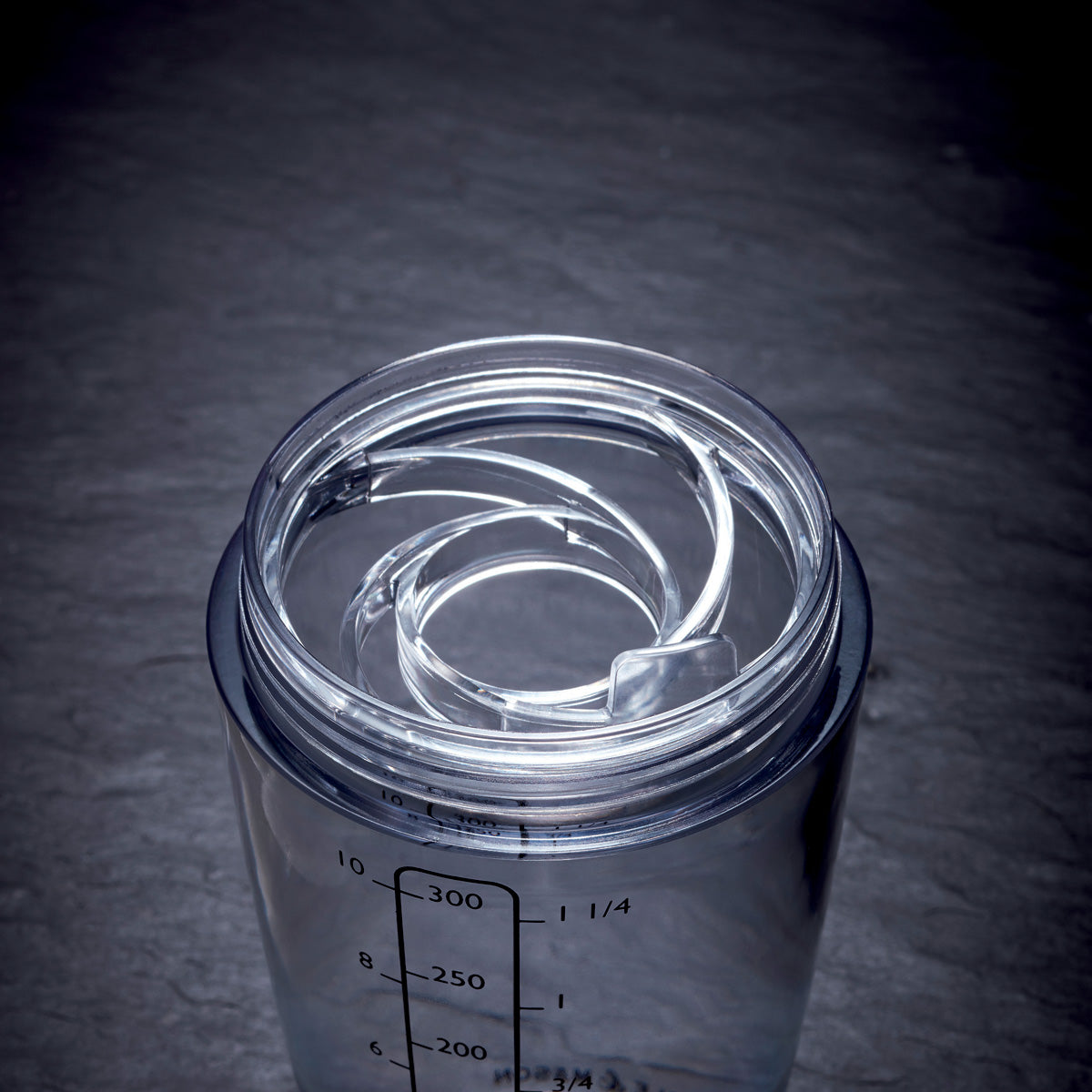 A close-up of a transparent plastic cup with measurement markings and a spiral-shaped clear straw inside, placed on a dark, textured surface.