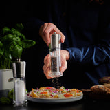 Someone grinds black pepper with the Cole & Mason Sandown Salt & Pepper Mill Set, featuring adjustable grinding for precise seasoning. A salt grinder, fresh basil, and bread sit nearby on the table against a dark backdrop.