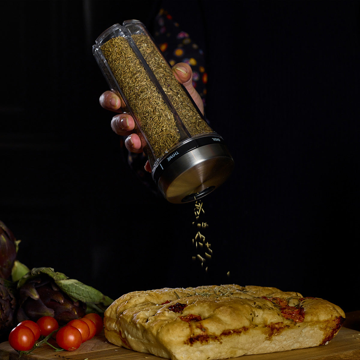 A hand shakes dried herbs from a transparent spice grinder onto a loaf of bread on a wooden surface. Cherry tomatoes and leafy vegetables are nearby. The background is dark.