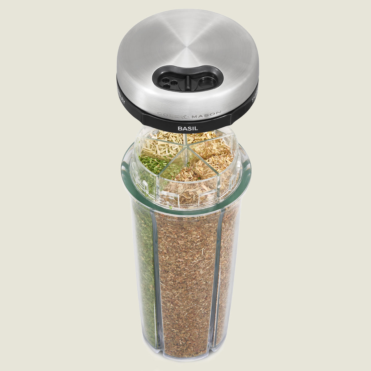 A clear glass spice jar with a stainless steel lid, divided into four sections containing different dried herbs. Each section is labeled, and the label Basil is visible on the rotating black band below the lid.