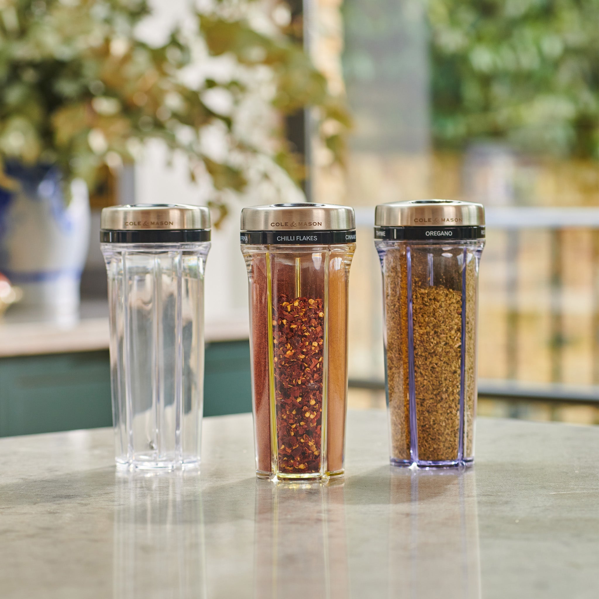 Three clear spice containers with silver lids labeled Salt, Chilli Flakes, and Oregano are arranged in a row on a kitchen counter, with a blurred background of plants and windows.