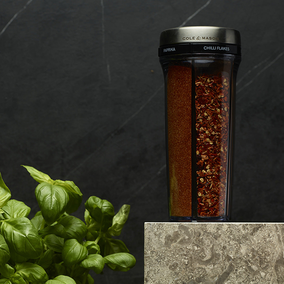 A dual spice grinder containing paprika and chili flakes sits on a gray marble surface, with fresh basil leaves nearby against a dark background.
