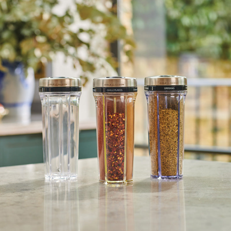 Three clear spice jars with silver lids are on a kitchen counter. One jar is empty, one contains chili flakes, and one contains oregano. Blurred greenery and a window are in the background.