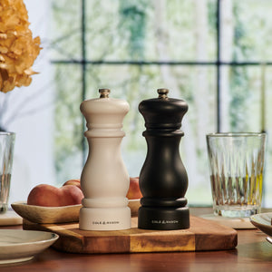 Two Cole & Mason Southwold Capstan Salt & Pepper Mills, in black and white, rest together on a wooden board. Their durable build and adjustable grind offer ideal seasoning among peaches, dishes, and sunlit windows.