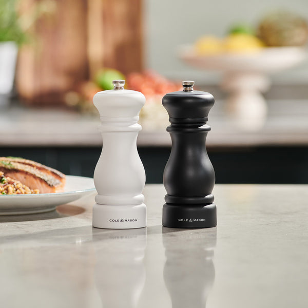 A white and a black Cole & Mason Southwold Capstan Salt & Pepper Mill set stand side by side on a kitchen counter, featuring adjustable grinding, with blurred food and kitchen items in the background.