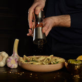 A Cole & Mason Stadhampton Chilli & Spice Mill grinds red pepper flakes over pasta, while fresh garlic, an olive-filled bowl, and a lemon wedge rest on a dark table nearby.