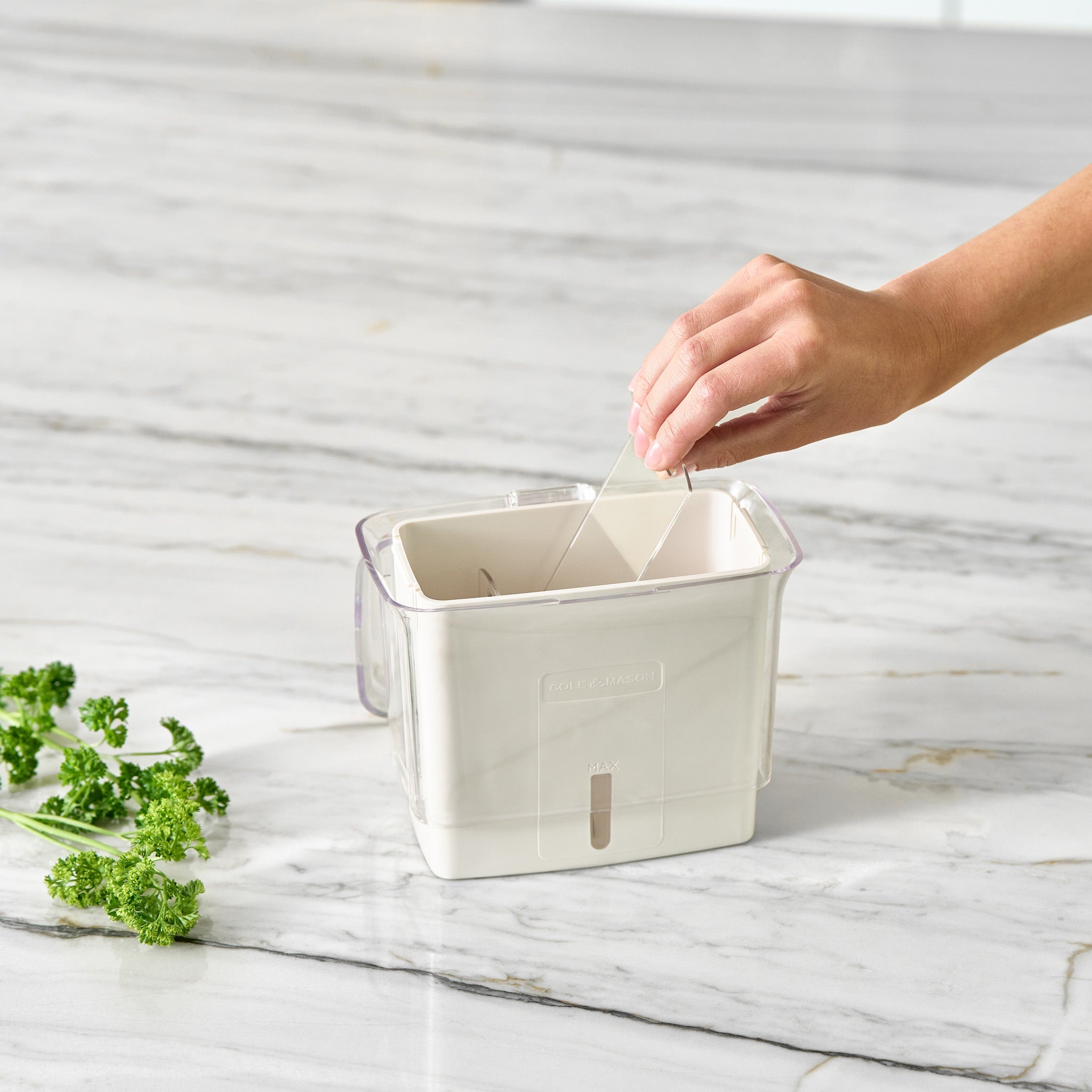 A hand pulls a string attached to a white plastic kitchen tool on a marble countertop, with a few sprigs of curly parsley nearby.