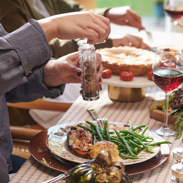 At a festively set table, someone uses the Cole & Mason Wallis Professional Nutmeg Grinder—with its ergonomic design—to grind pepper onto green beans, meat, and a roll while another diner enjoys wine and pie in the background.