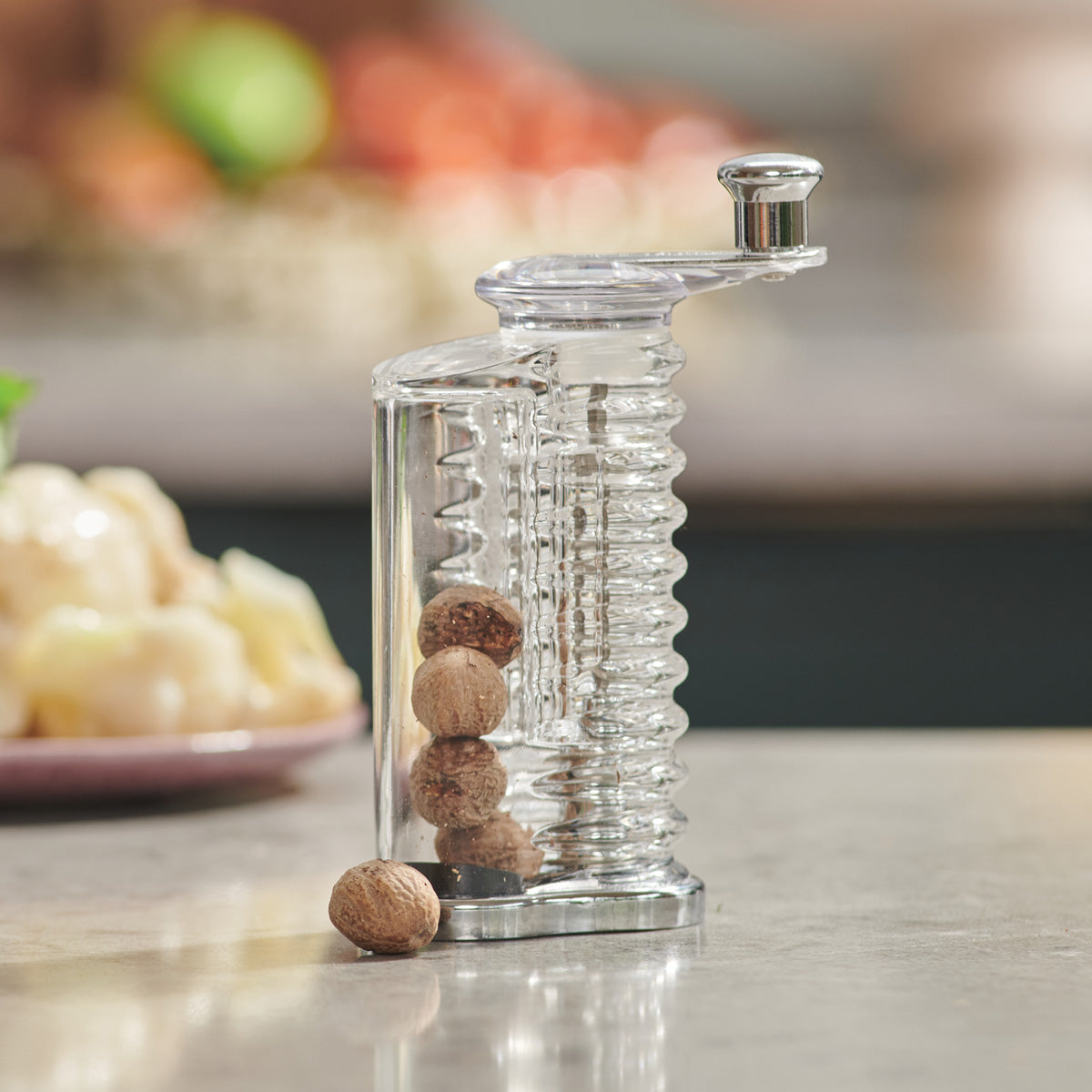 A clear plastic nutmeg grinder with several whole nutmeg seeds inside sits on a light kitchen countertop, with blurred food and kitchen items in the background.