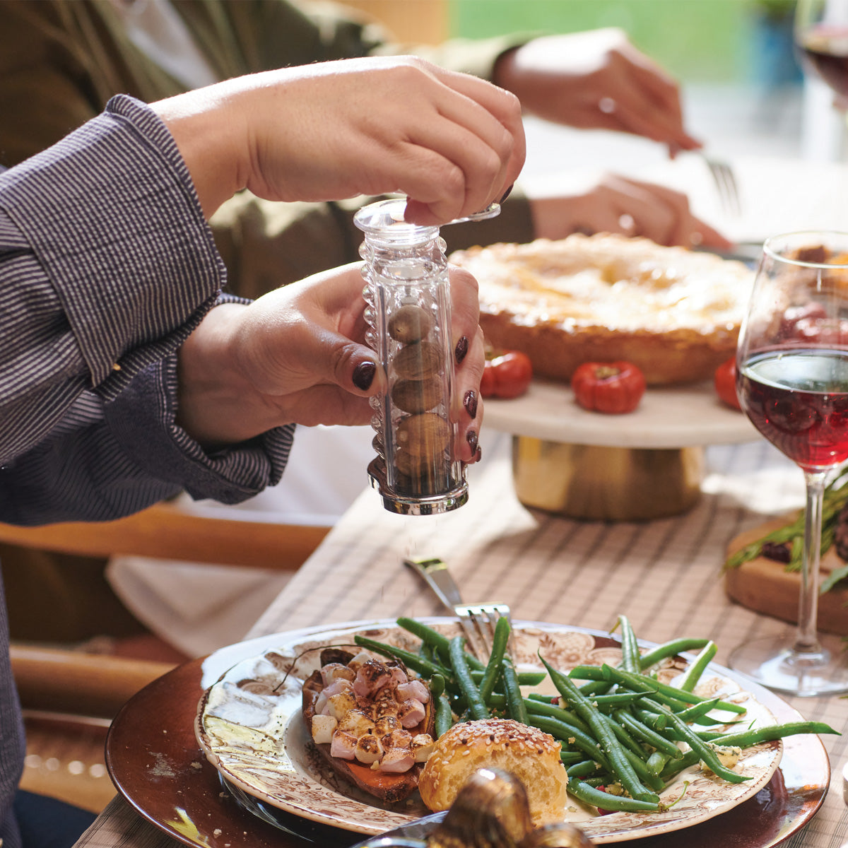 A person grinds pepper onto a plate of green beans, roasted potatoes, and meat at a dining table with a glass of red wine and a pie in the background.