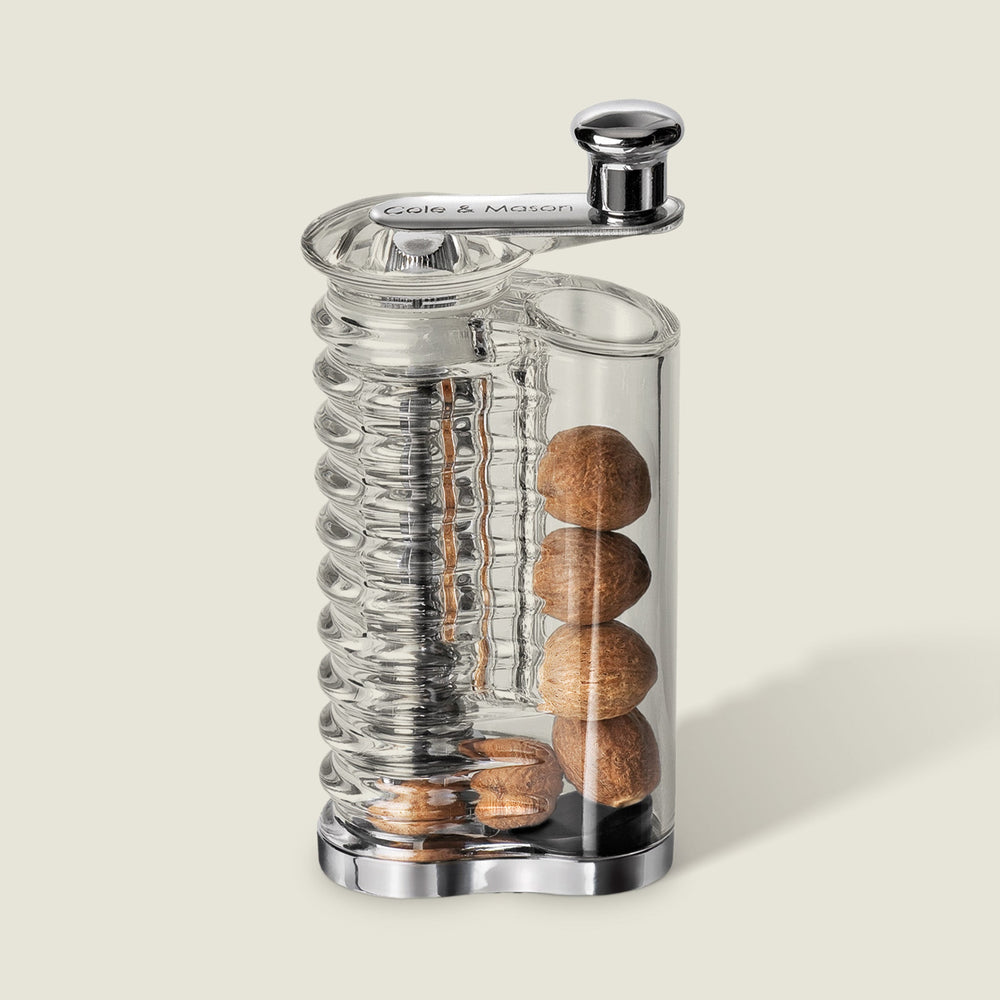The Cole & Mason Wallis Professional Nutmeg Grinder is a clear glass grinder with an ergonomic design, stainless steel blade, metal crank handle, vertical spiral mechanism, and side storage for whole nutmeg.