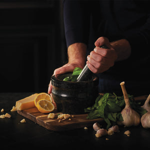 #140mm

A person uses a mortar and pestle to grind fresh basil. Nearby, garlic bulbs, lemon slices, Parmesan cheese, and chopped garlic rest on a wooden cutting board. The scene is dimly lit, emphasizing the ingredients.