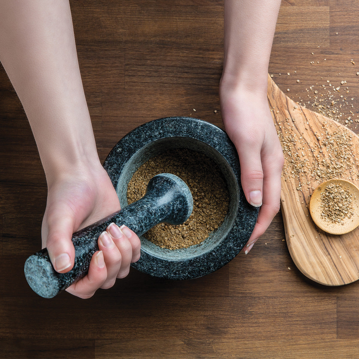 A person uses a black stone mortar and pestle to grind spices on a wooden surface, with some ground spices and a small wooden spoon on a nearby cutting board.