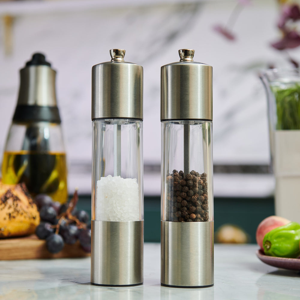 The Cole & Mason Everyday Stainless Steel Salt & Pepper Mill Set features an ergonomic design and adjustable coarseness; both mills stand upright on a light background, one containing coarse salt, the other whole black peppercorns.
