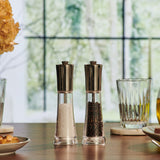 The Cole & Mason Everyday Style Salt & Pepper Mill Set, with adjustable grinders and metal tops, sits on a wooden table among plates, glassware, and a yellow flower; greenery is visible through the window behind them.