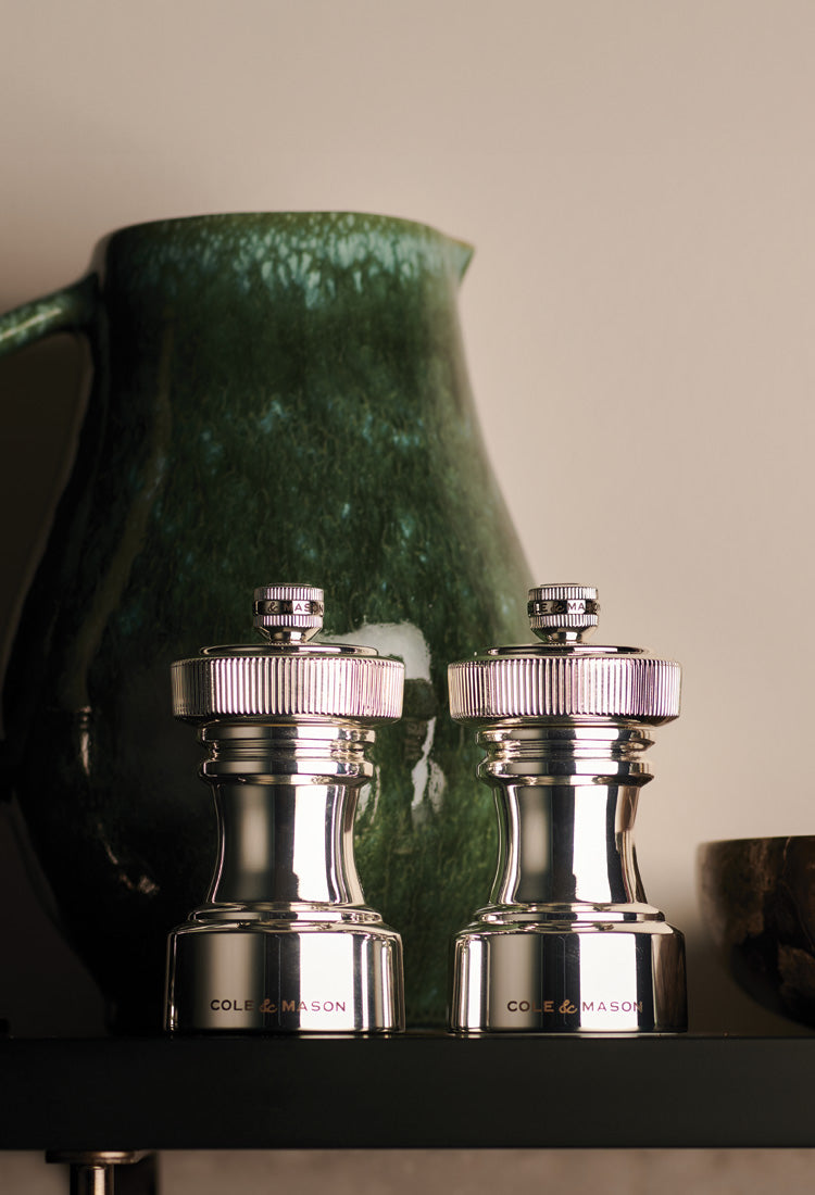 Two shiny metal salt and pepper grinders labeled Cole & Mason are placed side by side on a shelf, with a large, textured green ceramic pitcher in the background.